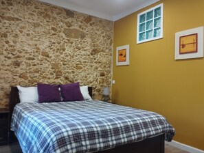 Un dormitori amb paret de pedra, un llit amb edredó escocès, una nitzeta, una làmpada i dos quadres al mur.