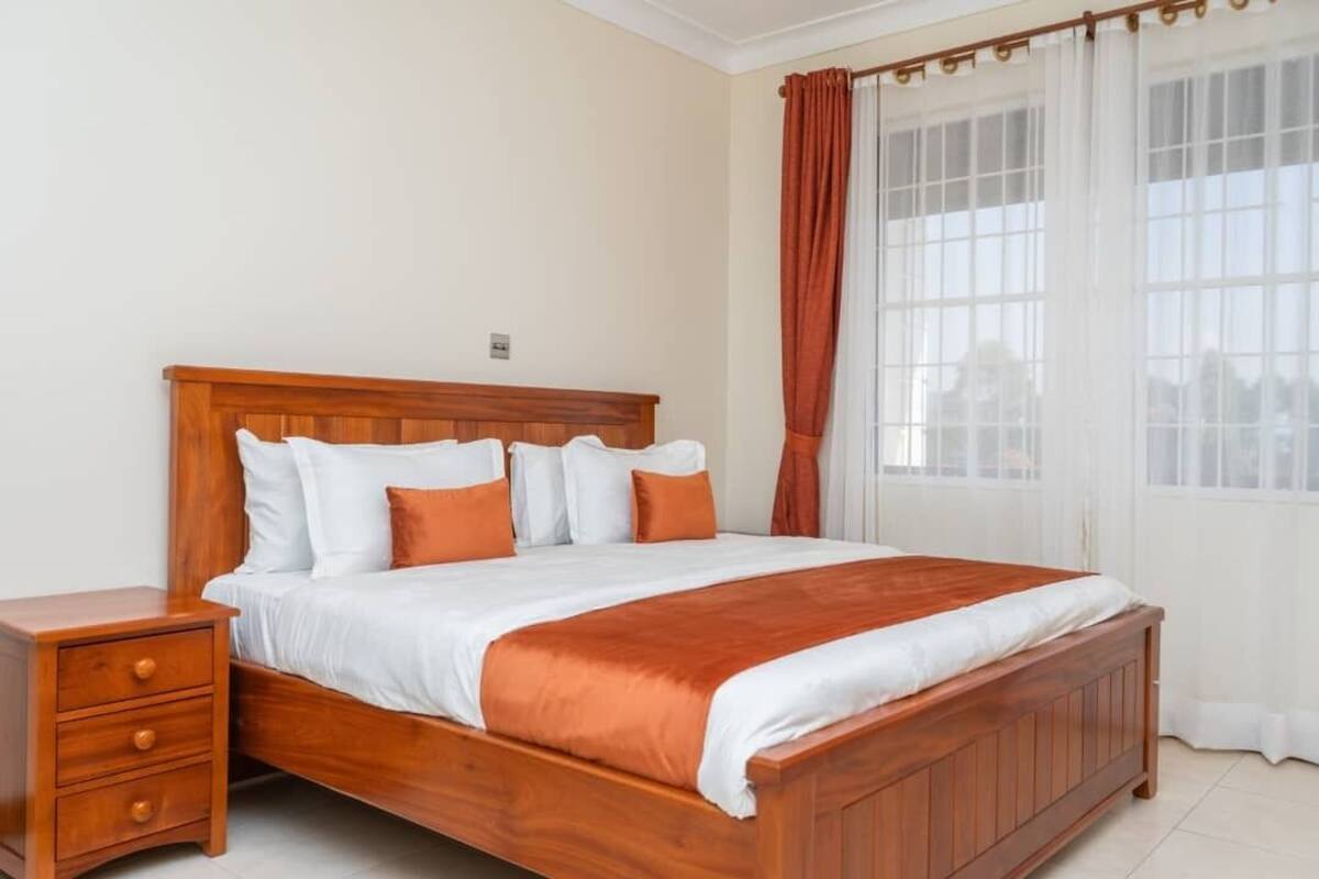 A wooden bed with white and orange bedding, a nightstand with drawers, and a window with curtains.