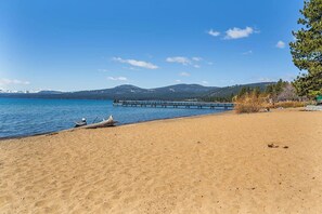 A beach with a wooden pier extending into calm waters, surrounded by mountains and sparse vegetation.