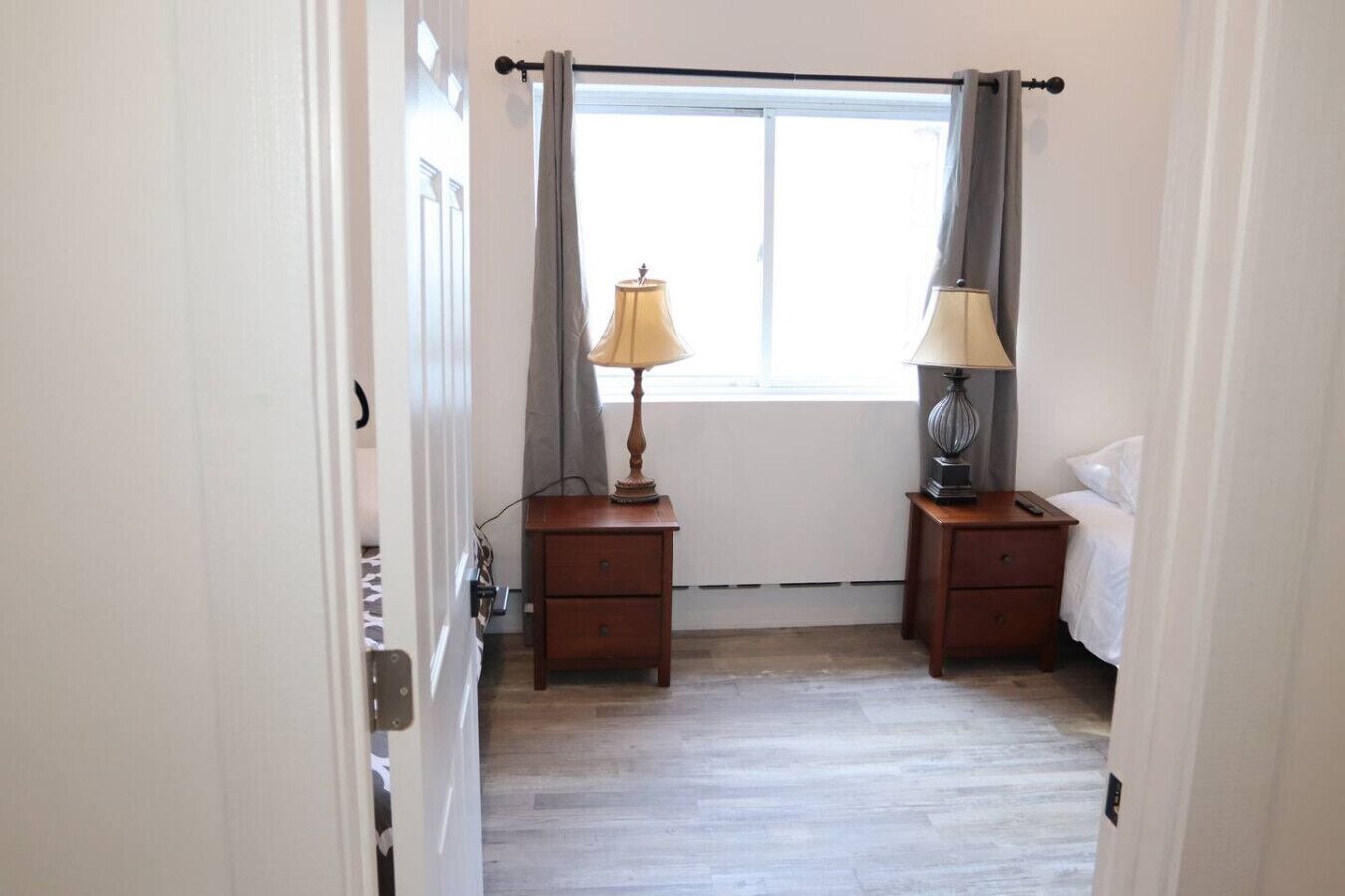 A bedroom with a bed, two bedside tables with lamps, a window with curtains, and a wooden floor.