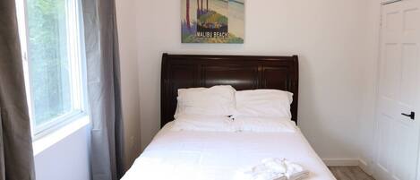 A neatly made bed with white linens, a dark wooden headboard, a window with curtains, and a wall-mounted picture of Malibu Beach.