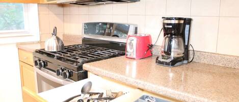 Una cocina con estufa, tostadora, cafetera y cajón con utensilios.