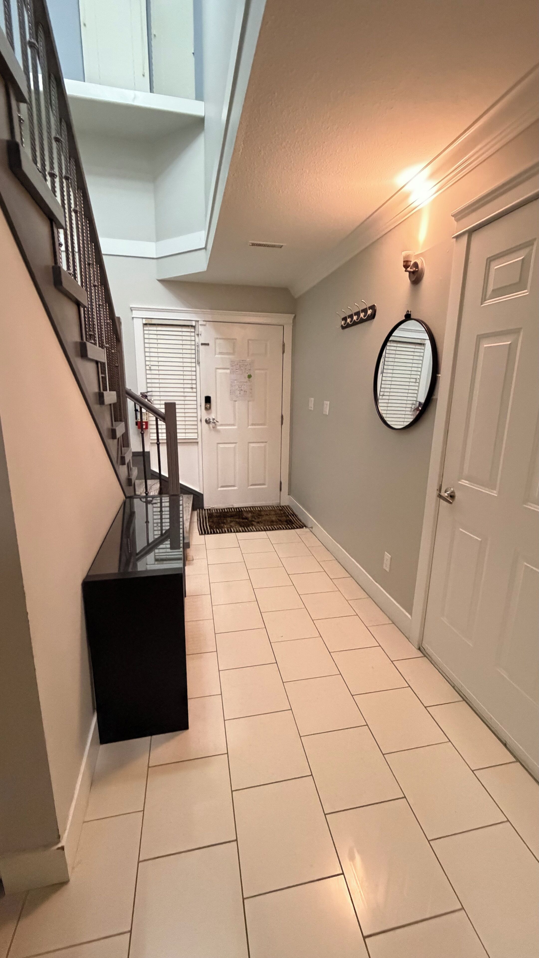 A hallway with a staircase, a door with a mat, a coat rack, and a wall-mounted mirror.