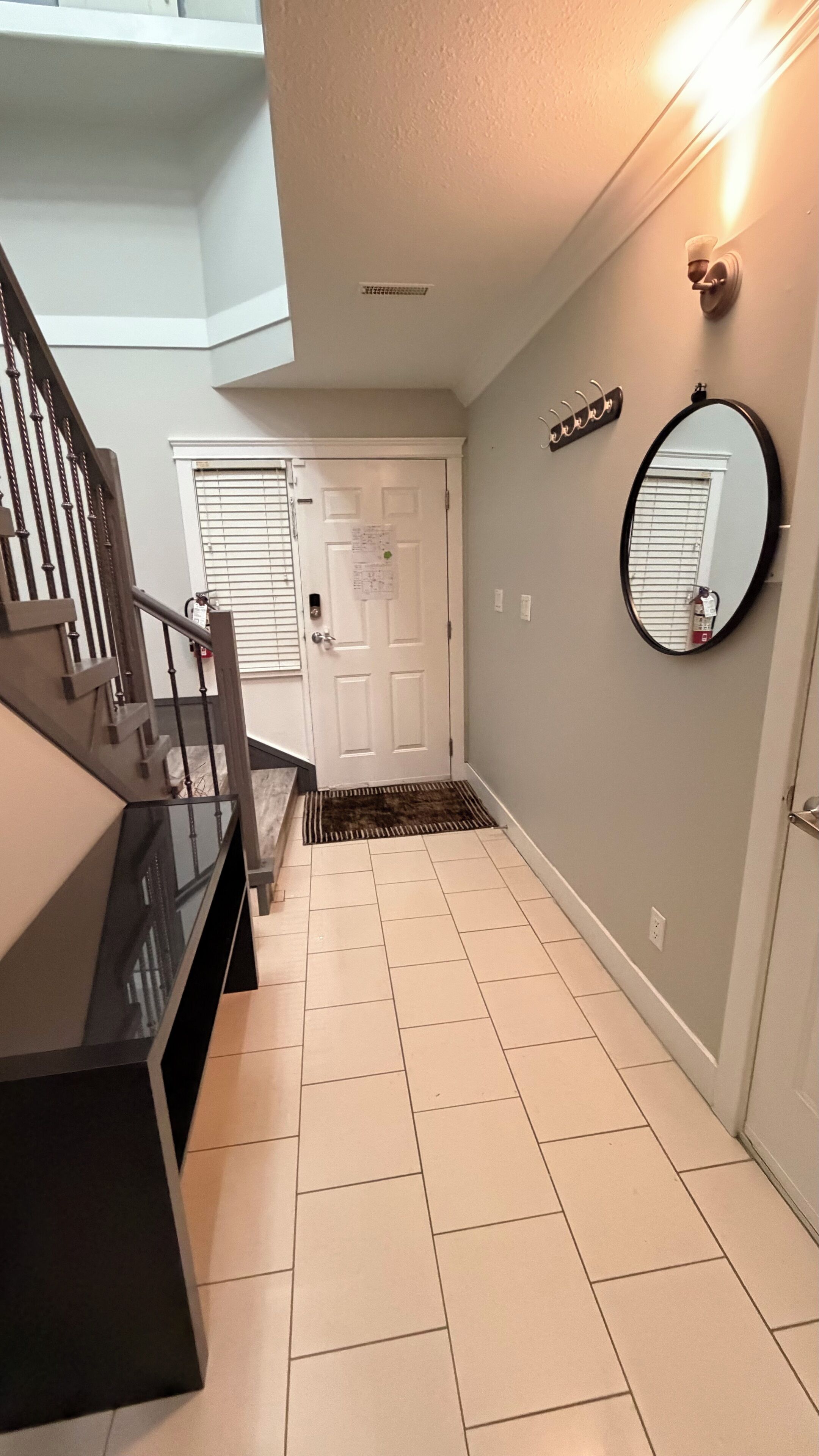 Un couloir avec un escalier, une porte blanche, un miroir et un luminaire fixé au mur.
