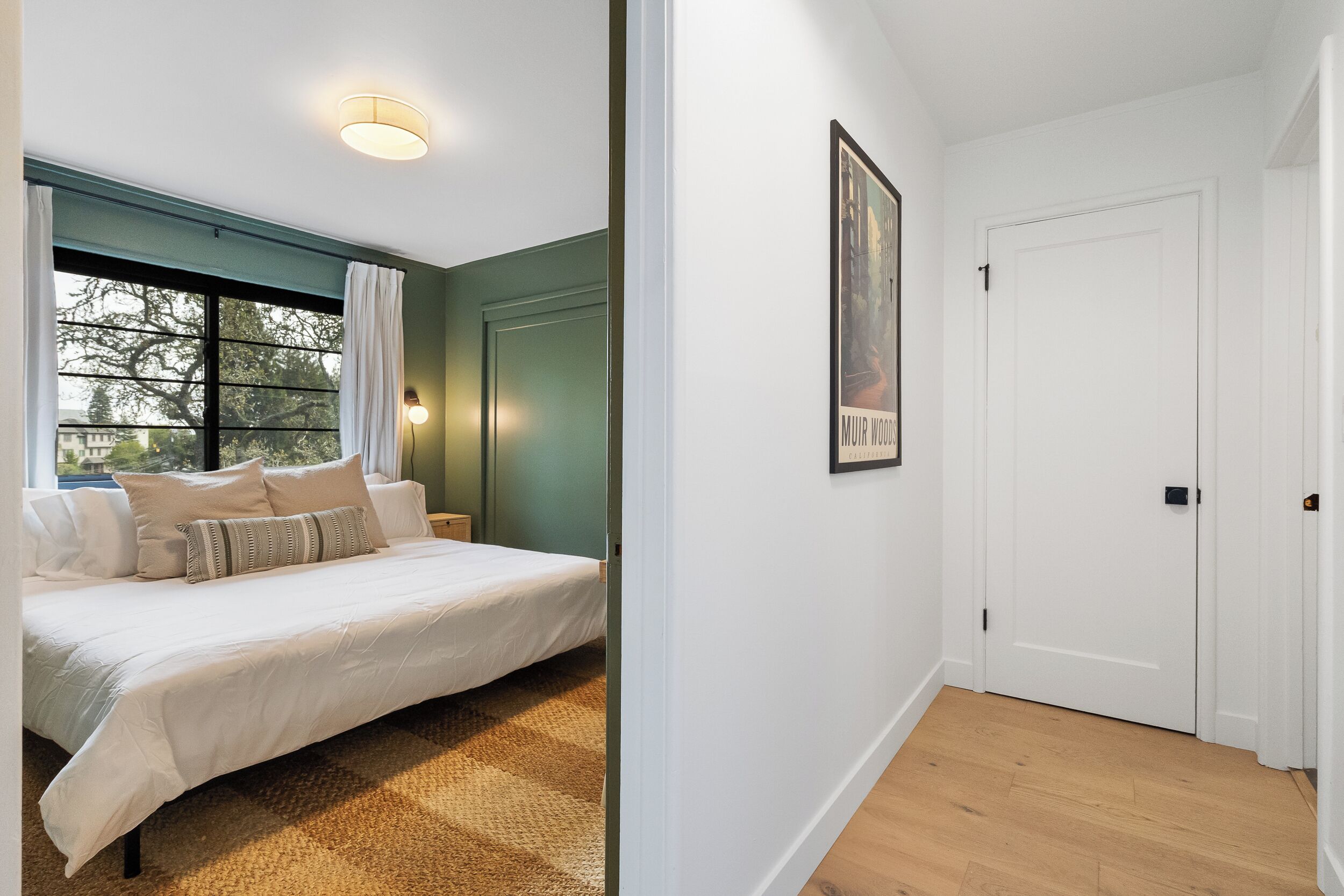 Un dormitorio con cama, ventana con cortinas, luz empotrada en la pared y una puerta.