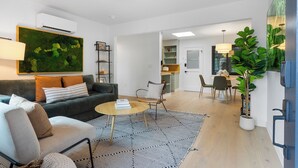 Un salon moderne avec un canapé, une table basse et des fauteuils, un coin repas avec une table et des chaises, et une cuisine en arrière-plan.