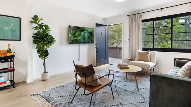 Un salon moderne avec un canapé, une table basse et un fauteuil. Il y a une grande fenêtre avec des rideaux, une plante en pot et un téléviseur à écran plat fixé au mur.