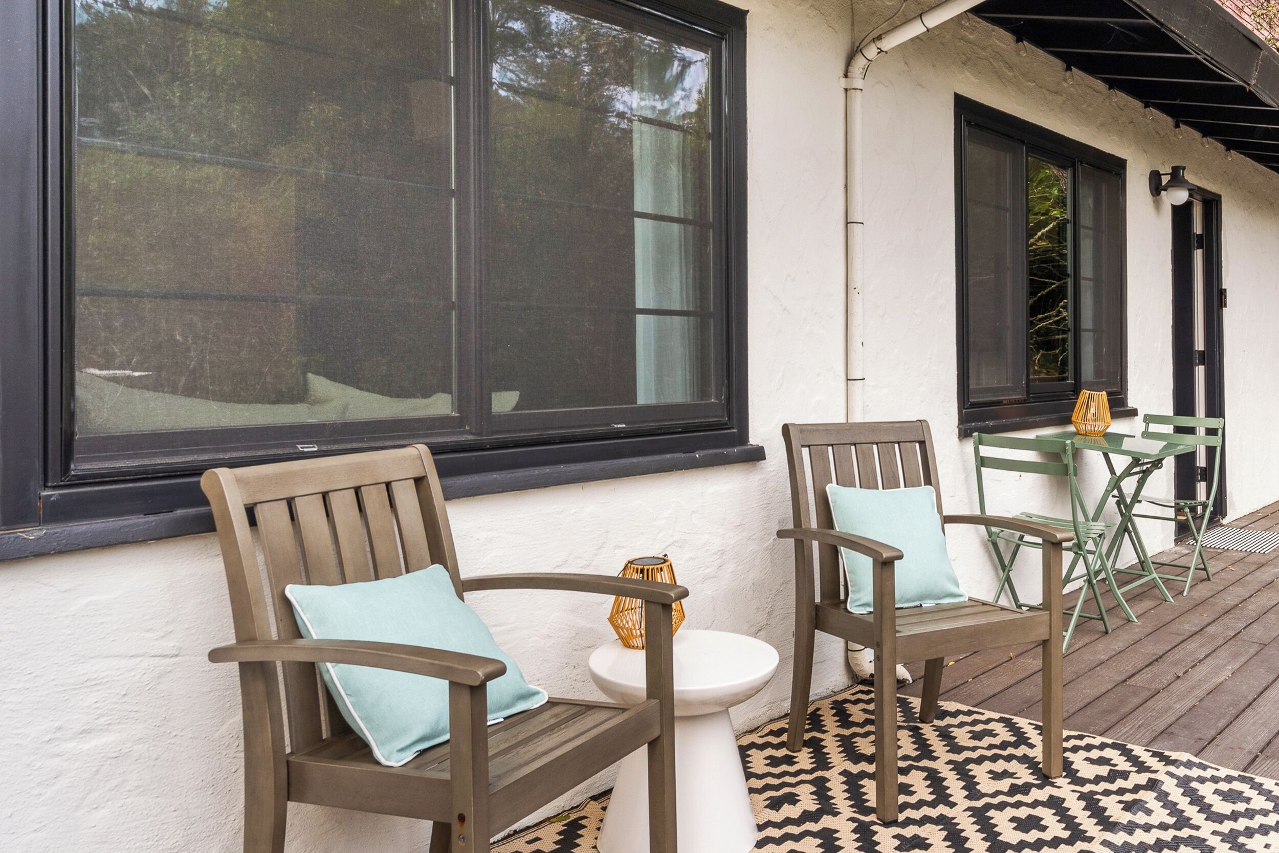 Eine Terrasse mit zwei Holzstühlen, einem kleinen weißen Tisch und einem gemusterten Teppich.