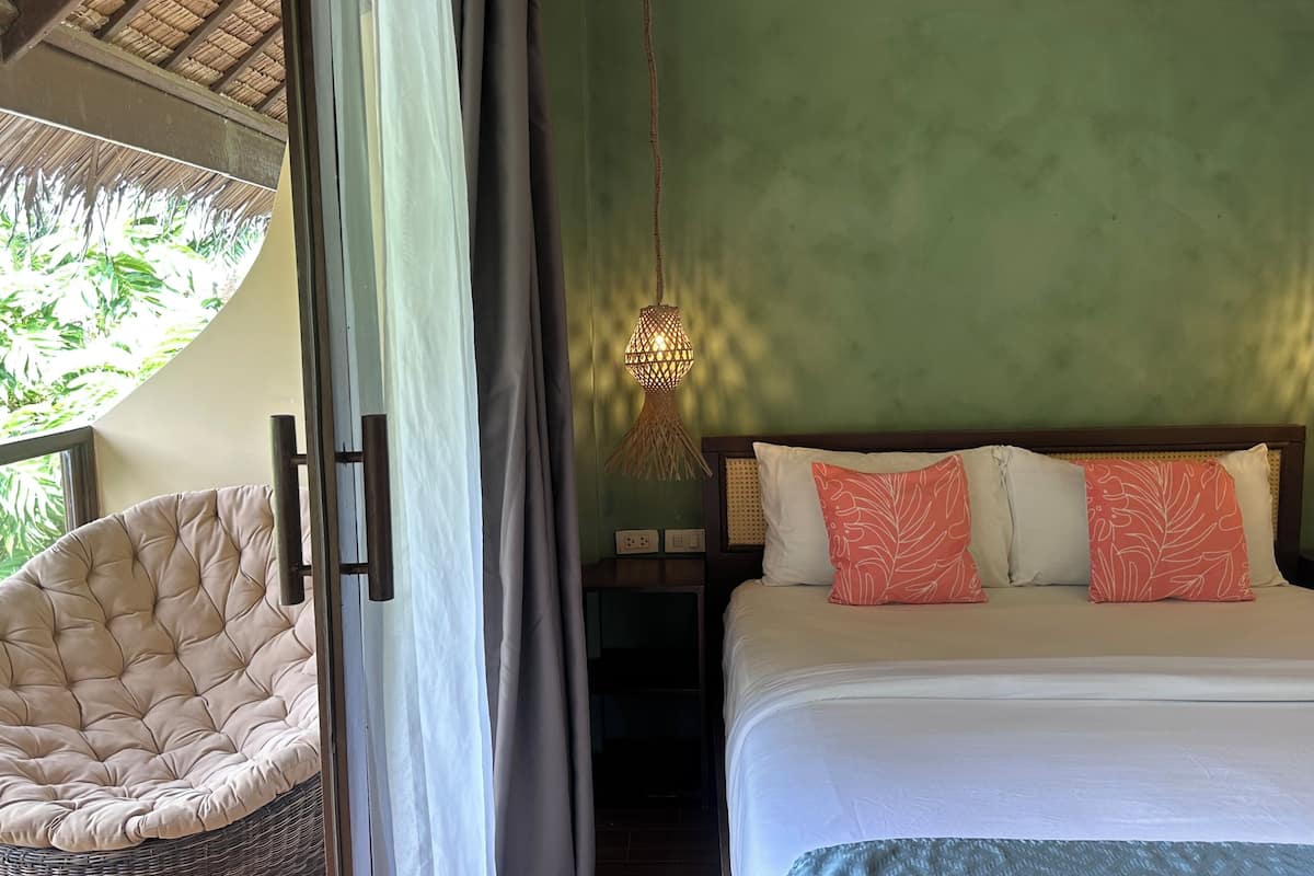 A bedroom with a bed, a hanging light fixture, a chair, and a view of greenery outside.