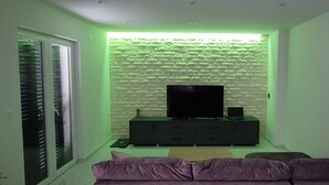 Ein modernes Wohnzimmer mit einem Flachbildfernseher auf einer schwarzen Unterhaltungsecke, indirekter grüner Beleuchtung und einem violetten Sofa.