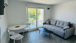 A modern living room with a grey sofa, a round dining table with chairs, and a large sliding glass door leading to an outdoor area. - Stylish Couple Getaway Near Beach (Sukosan)