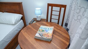 A neatly made bed with white linens, a bedside table with a lamp, and a round wooden table with a seafood cookbook. - Amila Resort (Habarana)