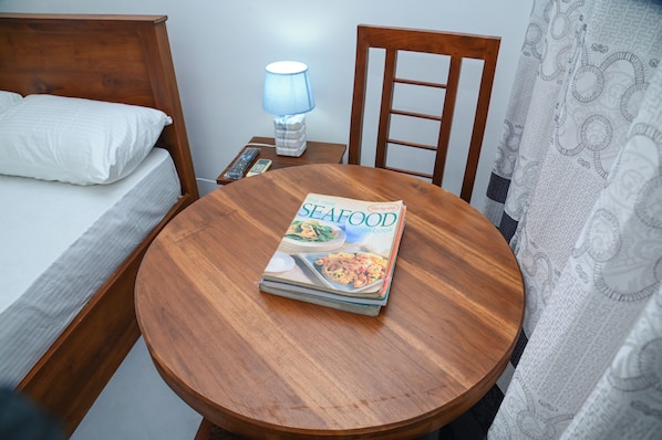 A neatly made bed with white linens, a bedside table with a lamp, and a round wooden table with a seafood cookbook.