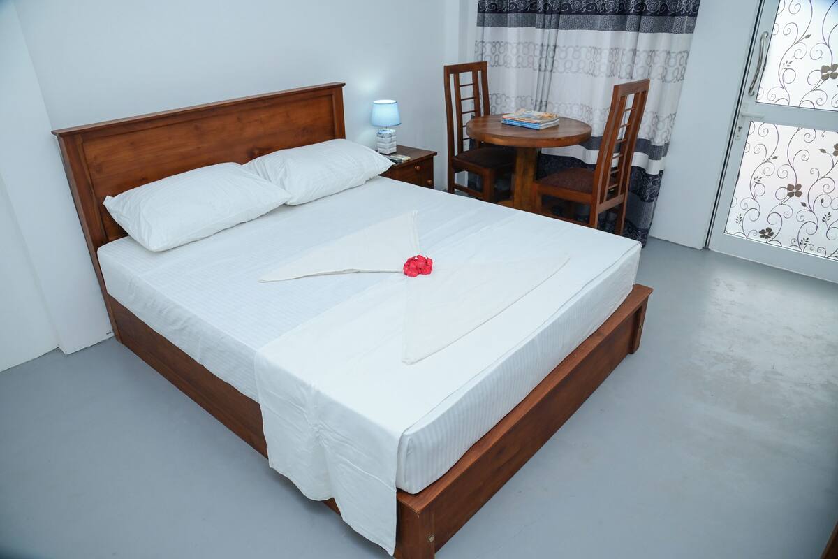 Una cama bien tendida con sábanas blancas, cabecera de madera, mesita de noche con lámpara, una silla y una pequeña mesa con un libro y una flor.