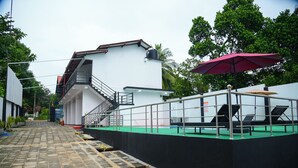 A modern building with a staircase leading to a balcony, a covered seating area with umbrellas, and a paved pathway. - Amila Resort (Habarana)