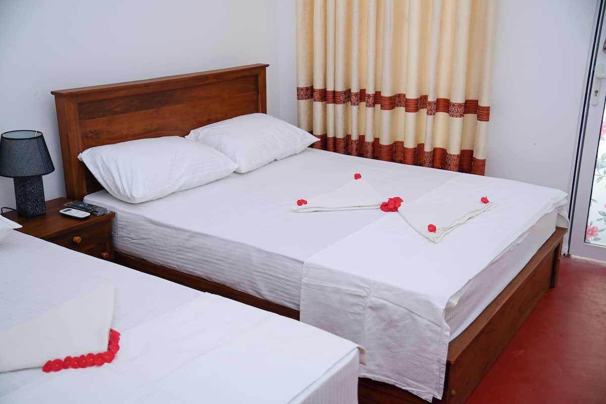 Una cama doble con ropa de cama blanca y flores rojas, un cabecero de madera, una mesita de noche con una lámpara y una ventana con cortinas.