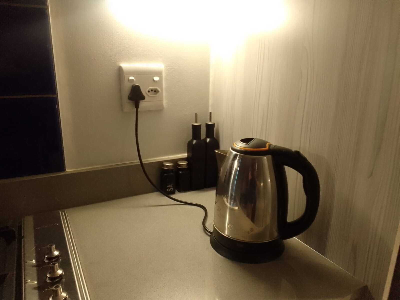 Una tetera de acero inoxidable sobre una encimera blanca, con un enchufe y dos botellas al fondo.