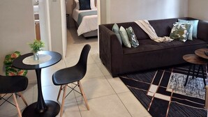 A modern living room with a sofa, coffee table, and chairs. There is a bed in the background and artwork on the walls. - Oak & Olive Retreat Midrand (Midrand)