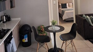 A compact living space with a kitchenette, a dining area with two chairs, a small table, and a bed in the background. - Oak & Olive Retreat Midrand (Midrand)
