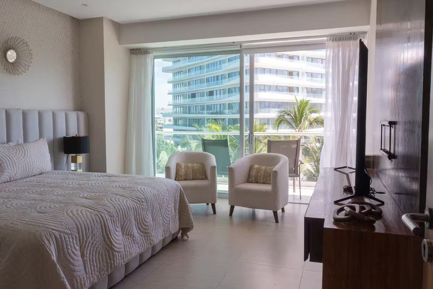 Ein Hotelzimmer mit einem großen Bett, zwei Sesseln, einem Fernseher und einem Balkon mit Blick auf ein Hochhaus.