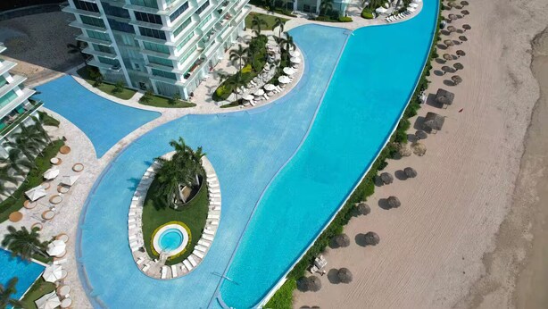 Eine Vogelperspektive zeigt einen Resort mit einem zentralen Schwimmbecken, einem Strandbereich und einem mehrstöckigen Gebäude mit Balkonen.