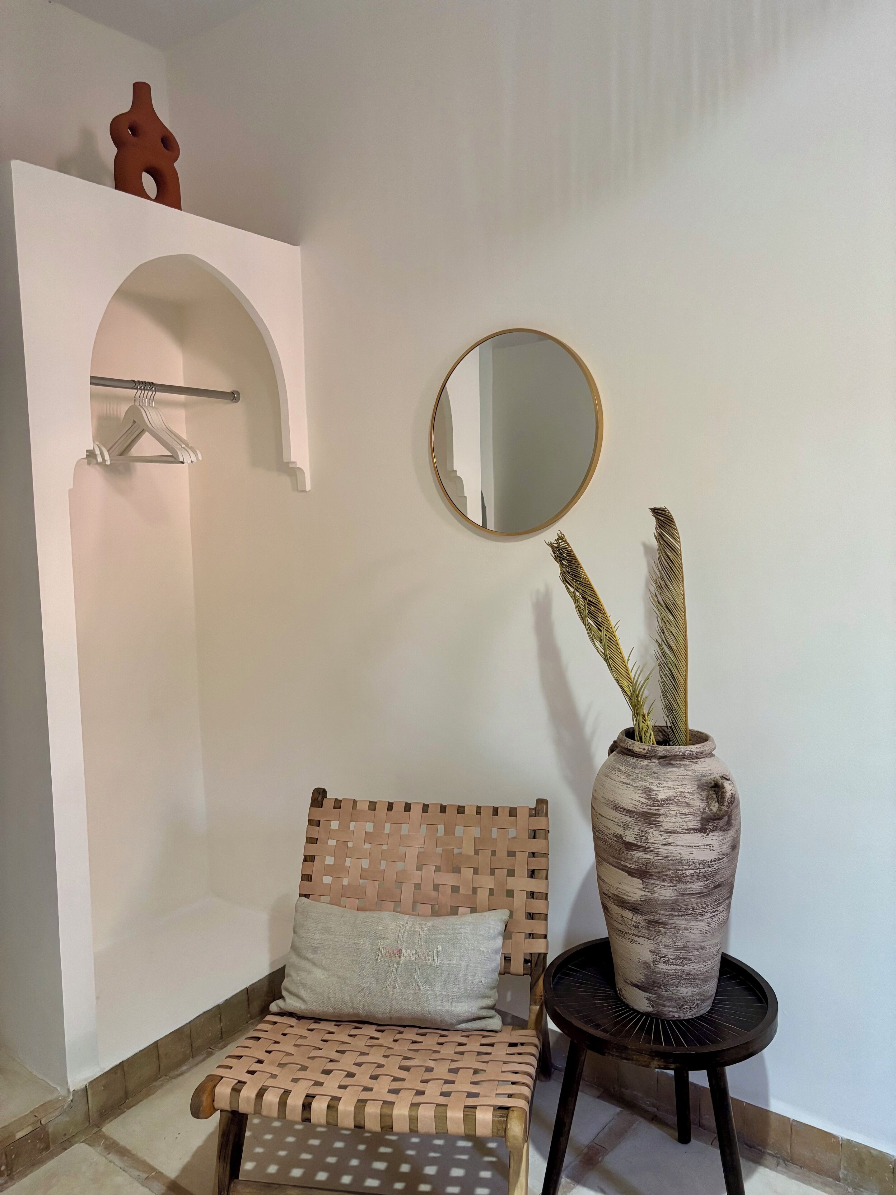 A room with a woven bench, a round mirror, and a vase with plants.