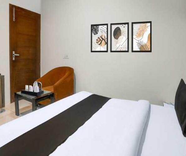 A hotel room with a bed, a chair, a small table with a kettle and cups, and three framed artworks on the wall.