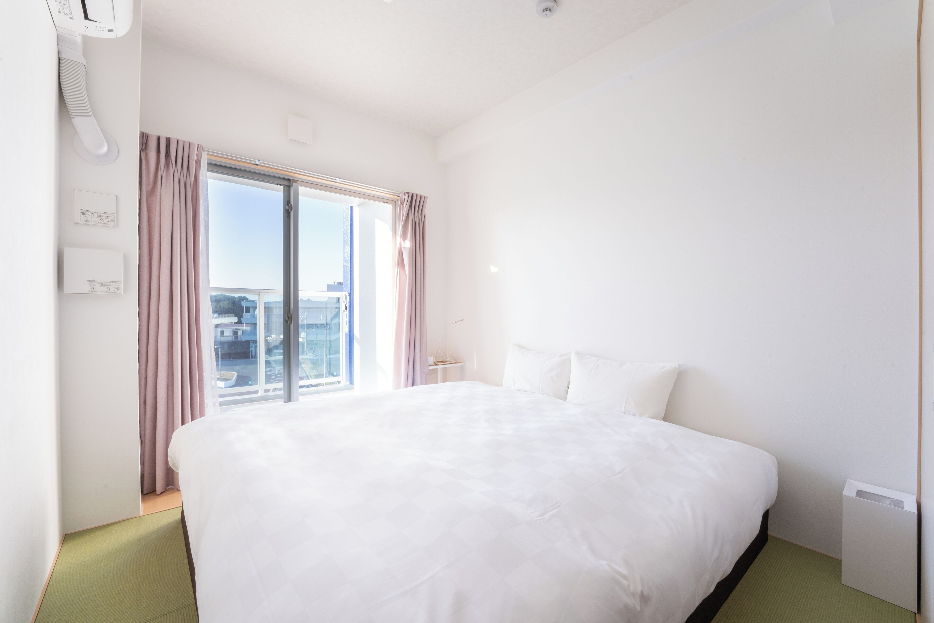 A hotel room with a large bed, a window with curtains, a bedside table, and a view of the city.