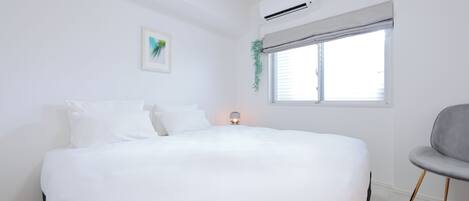 A bedroom with a large bed, a bedside table with a lamp, a window with blinds, and a chair.