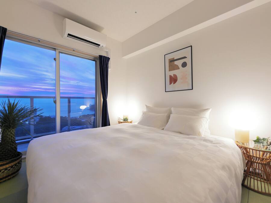 A hotel room with a large bed, a view of the sky, a wall-mounted artwork, and a small table with a lamp.