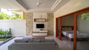 A modern room with a grey sofa, a flat-screen TV mounted on a stone wall, a bed with wooden headboard, and a sliding glass door leading to a balcony with greenery. - Juada Garden (Seminyak)