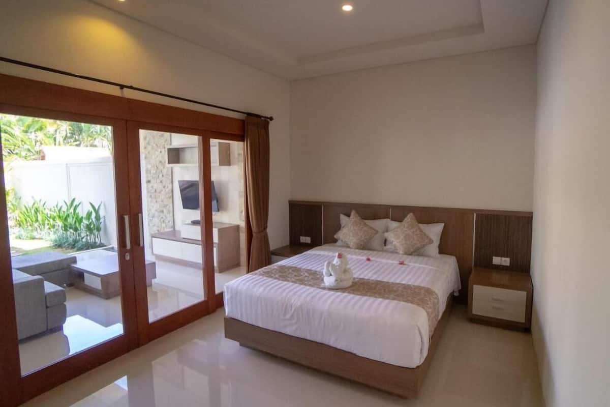 A modern bedroom with a large bed, a sliding glass door to a balcony, and a flat-screen TV on a stand.