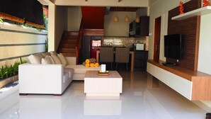 A modern living room with a sofa, a coffee table, and a TV. There is a staircase leading to a kitchen area. - Juada Garden (Seminyak)