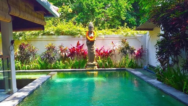 A swimming pool with a statue in the center, surrounded by lush greenery and a white wall. - Juada Garden (Seminyak)