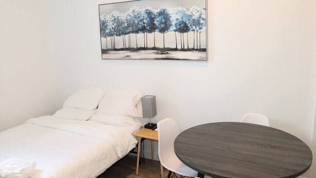 A single bed with white bedding, a bedside table with a lamp, a round table with chairs, and a painting of trees on the wall. - Budget 1BR for 2 Guests in Pavonia (Jersey City)