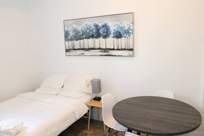 Una cama individual con ropa de cama blanca, una mesita de noche con lámpara, una mesa redonda con sillas y un cuadro de árboles en la pared.