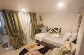 A bedroom with a bed, curtains, a nightstand, a vase with flowers, and a wall-mounted air conditioner. - Prime World District Condo (Lapu-Lapu)