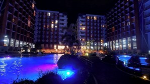 A nighttime view of a hotel with a swimming pool, illuminated by colorful lights, surrounded by modern buildings. - Prime World District Condo (Lapu-Lapu)