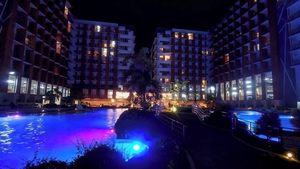 Ein Hotel mit Schwimmbad bei Nacht, beleuchtet durch farbige Lichter, umgeben von modernen Gebäuden.