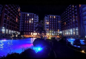 Ein Hotel mit Schwimmbad bei Nacht, beleuchtet durch farbige Lichter, umgeben von modernen Gebäuden.