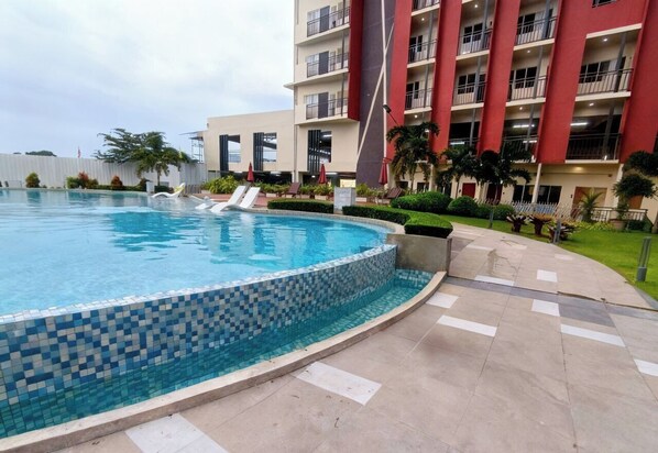 Une piscine dont le bord est recouvert de carreaux, entourée d’une allée pavée, et située à côté d’un immeuble à plusieurs étages, doté de balcons et d’une façade rouge.