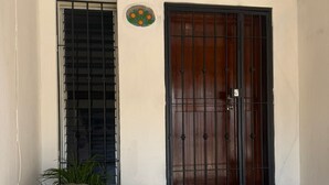 Une entrée d’immeuble avec un bac à fleurs en béton, des marches menant à une porte en bois surmontée d’un portail en métal, et un petit élément décoratif circulaire au-dessus de la porte.