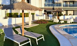 A poolside area with lounge chairs, a thatched umbrella, and a modern building in the background. - Apartments for 4 People (Orihuela)