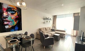 A modern living room with a sectional sofa, a dining table set for four, and a large abstract painting on the wall. - Apartments for 4 People (Orihuela)