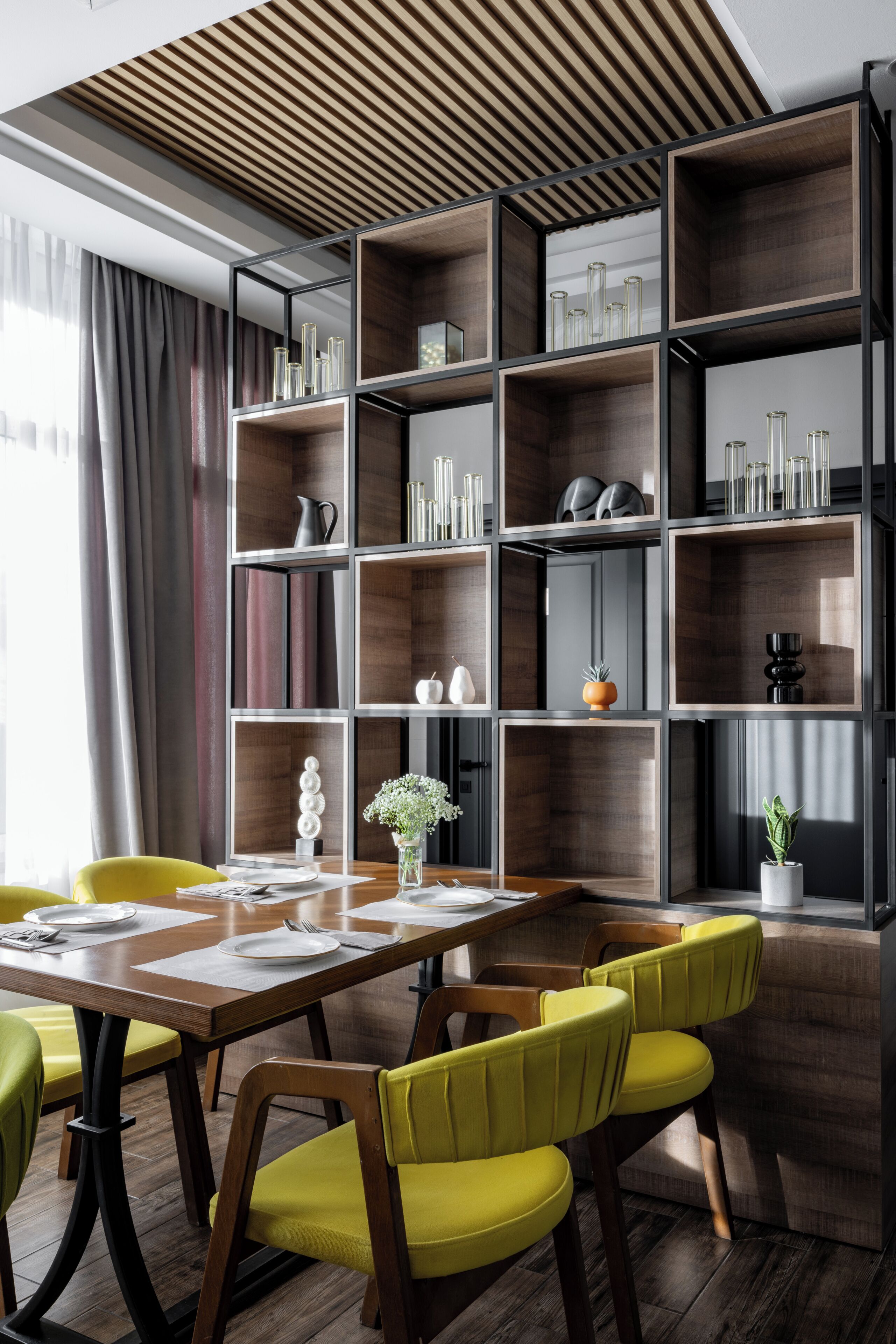 A modern dining area with a wooden table, yellow chairs, and a built-in bookshelf with various decorative items.