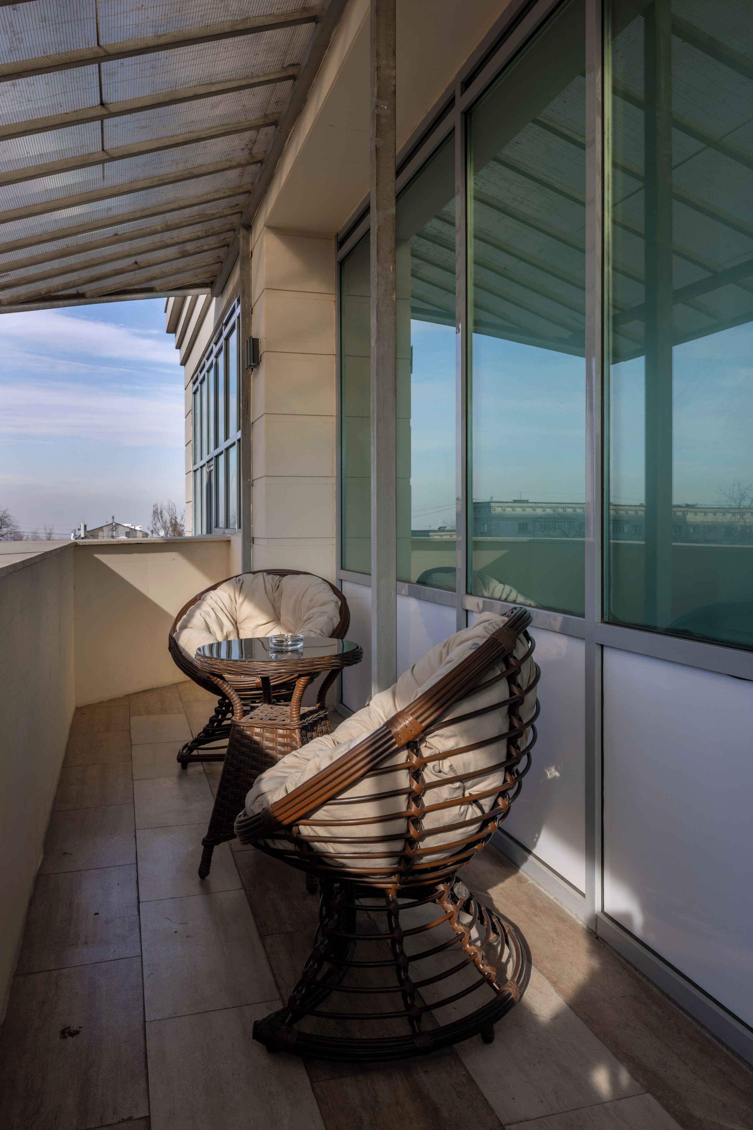 A balcony with wicker furniture, a glass table, and a view of the cityscape.