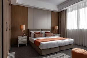 Una habitación de hotel moderna con una cama grande, dos mesitas de noche con lámparas, un armario y vistas al exterior a través de cortinas translúcidas.