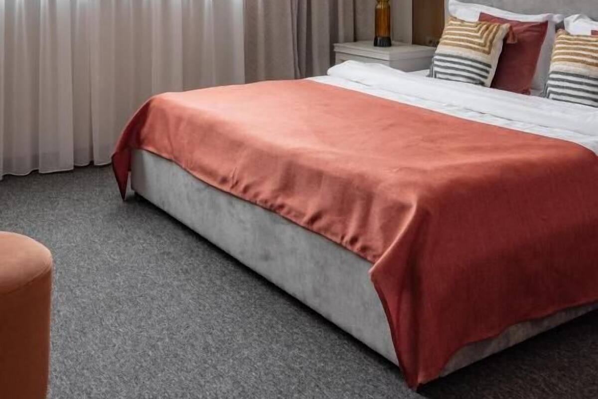 A hotel room with a large bed, a nightstand, a lamp, and a view of the outside through sheer curtains.