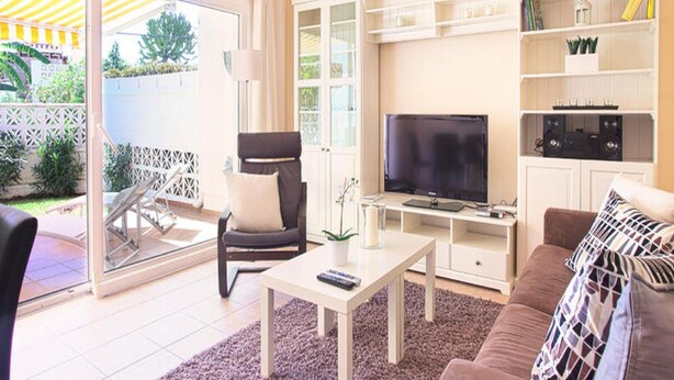 Ett vardagsrum med en tv, en soffa och ett soffbord. Där finns också en stol, ett glasbord och en bokhylla.
