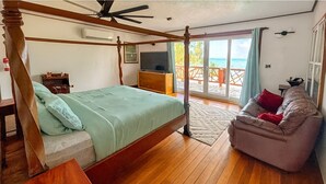 A bedroom with a wooden four-poster bed, a sofa, a TV, and a balcony with a view of the sea.